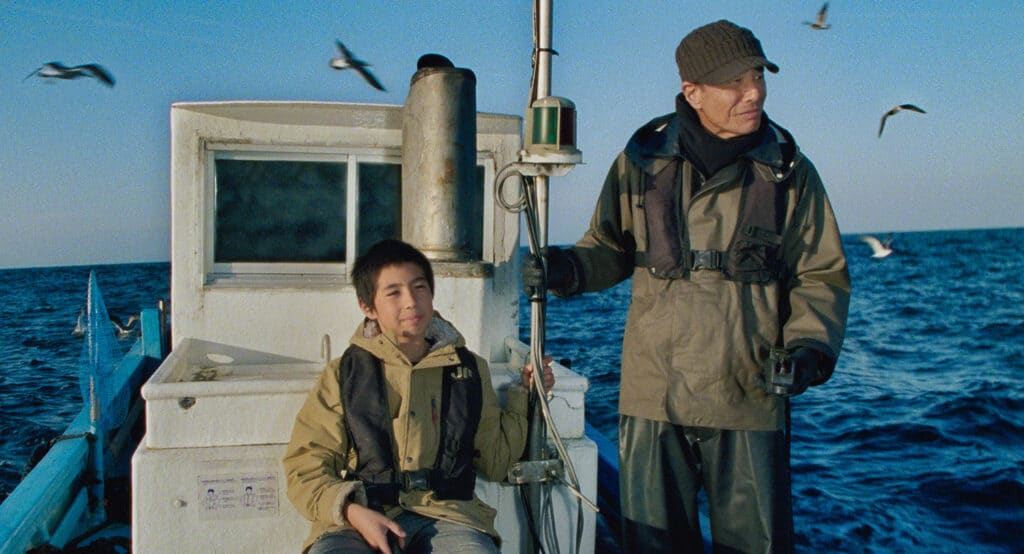 Scene from the 2025 Japanese film "港のひかり (Light of the Harbor)" featuring a young boy and older man on a fishing boat at sea, symbolizing a multigenerational bond.